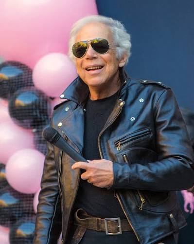 Ralph Lauren speaking into a microphone in a black leather jacket at the Pink Pony Walk launch event, in front of pink and black balloon arch