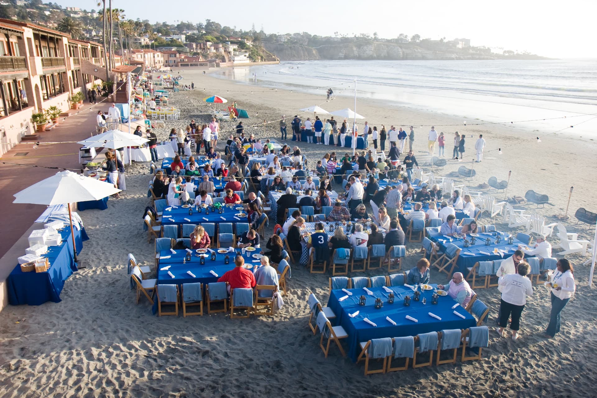Guests seated at blue-clothed tables on the beach at the La Jolla event
