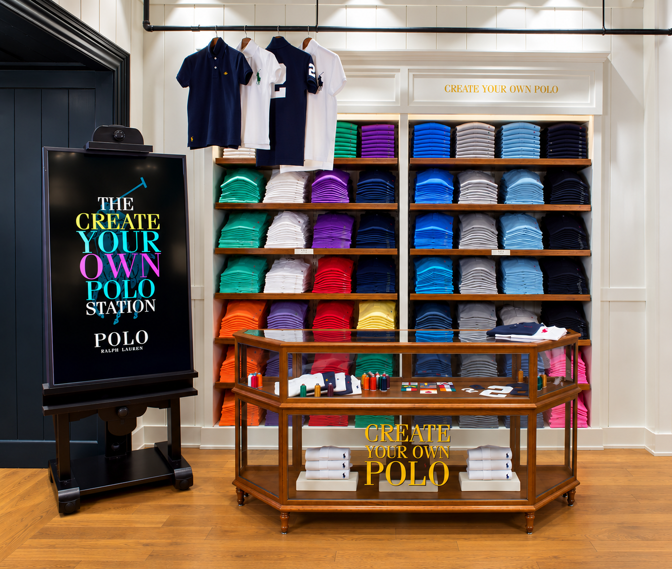Create Your Own Polo station with branded screen on an easel, wall of colorful folded polos and a wooden customization counter
