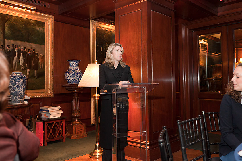 Speaker at a lectern beside a table lamp in the wood-paneled room