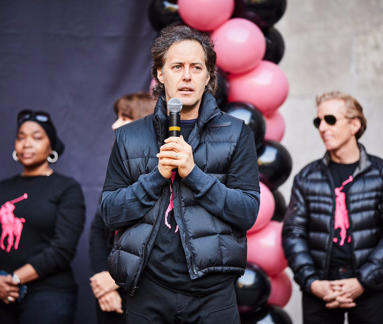 David Lauren speaking at a microphone at the Pink Pony Walk launch event, in front of pink and black balloon arch