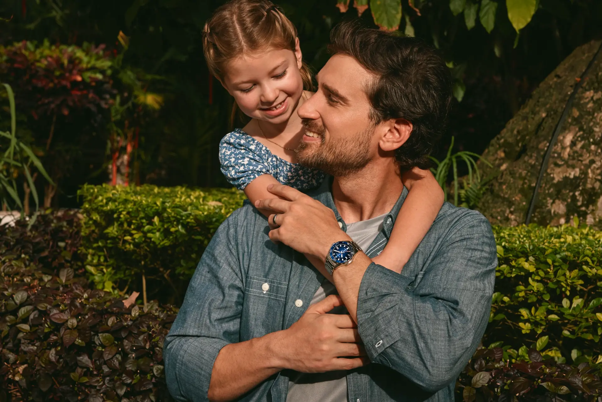 Lifestyle campaign photograph of a father wearing a blue-dial watch while his daughter hugs him from behind in a garden