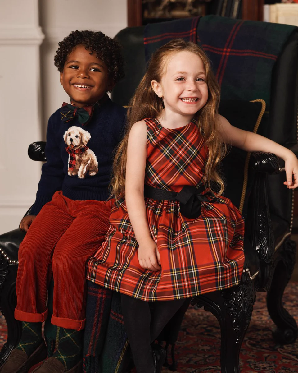 Two children seated in a dark leather wingback chair — a boy in a navy dog-intarsia sweater and red corduroy trousers and a girl in a red tartan dress