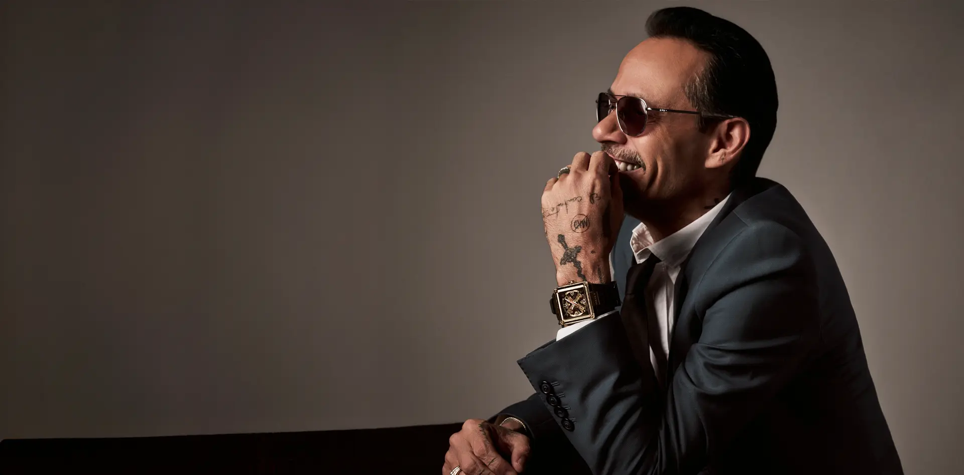 Marc Anthony wearing a Bulova watch in a campaign portrait, seated on the right against an open studio background