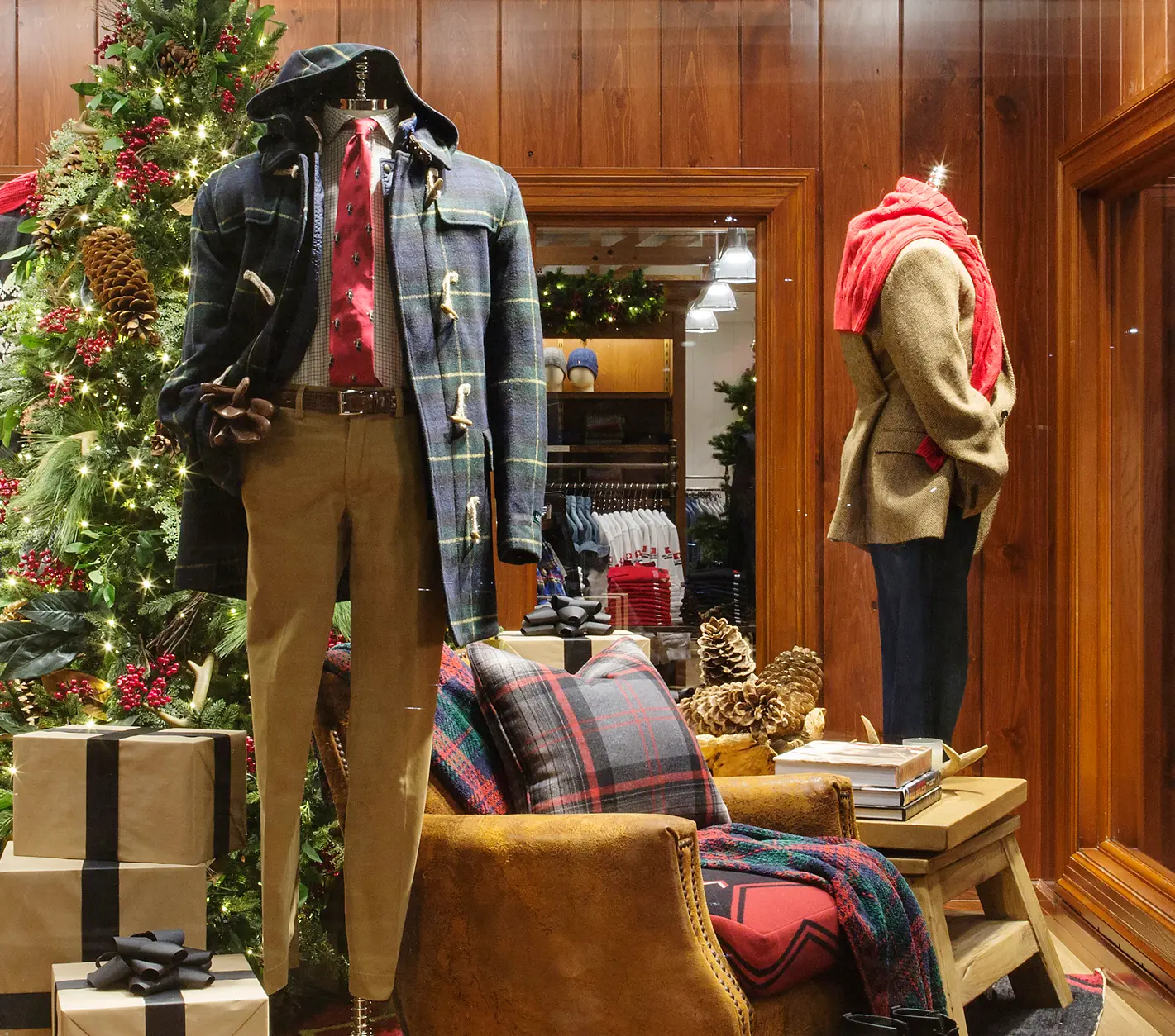 Ralph Lauren men's holiday environment with a male mannequin in a dark tartan duffle coat beside a lit Christmas tree, leather chair, tartan textiles, wrapped gifts and wood paneling