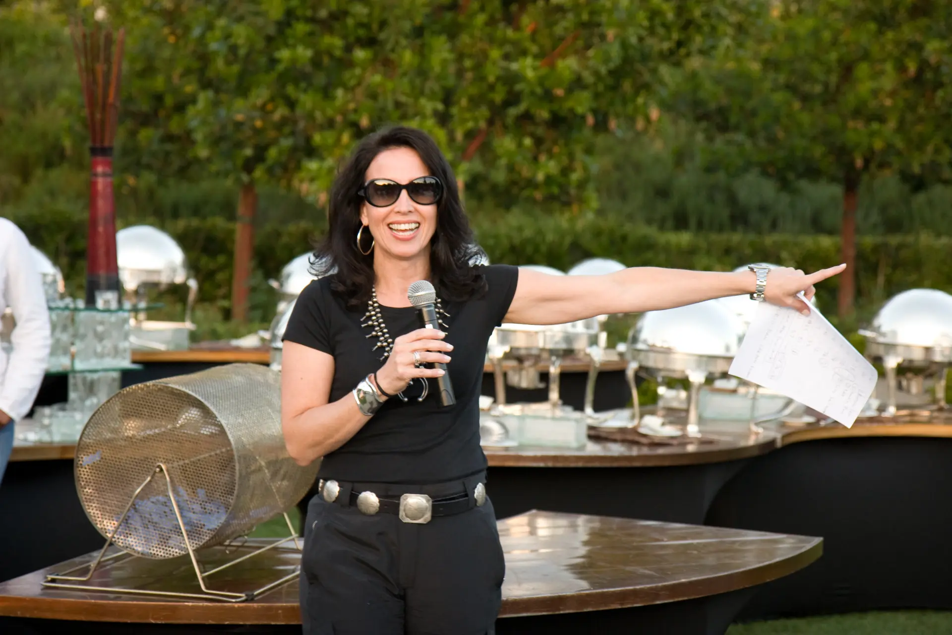 Tracey speaking into a microphone and pointing during the Las Vegas event, beside the raffle drum and catering setup