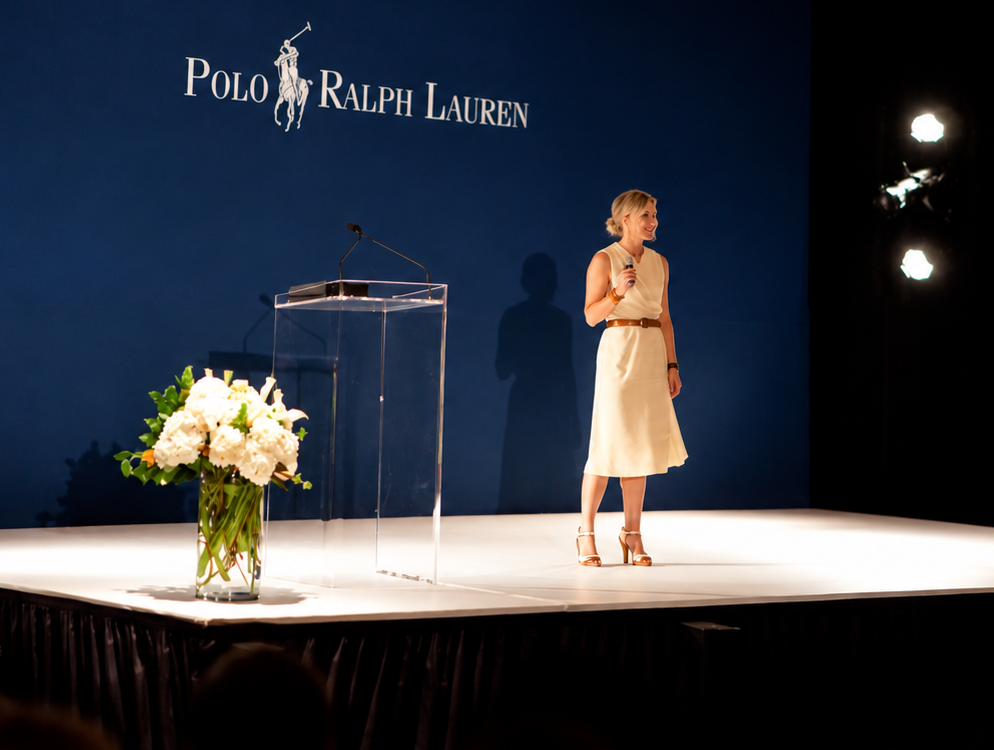 Speaker presenting on stage at the Polo Ralph Las Vegas conference, beside a clear podium with white florals