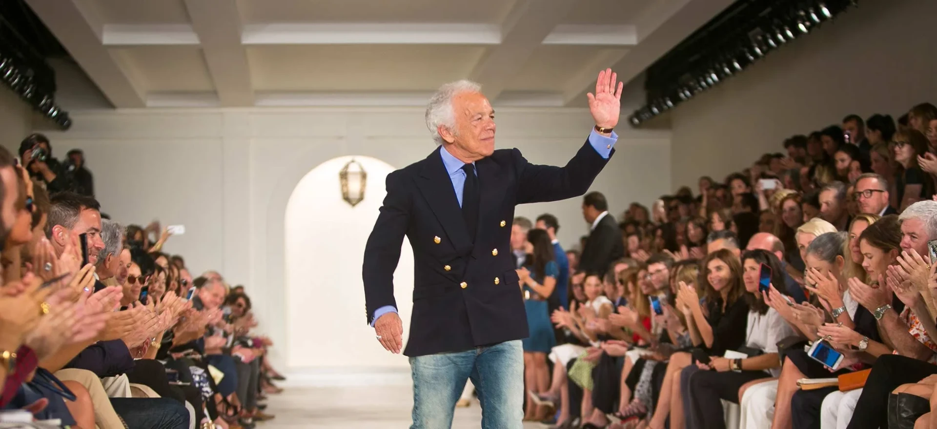 Ralph Lauren in a navy double-breasted blazer and jeans waving to the audience as he walks the runway